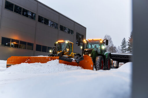 Brøytetraktorer med oransje skjær rydder snø ved et næringsbygg